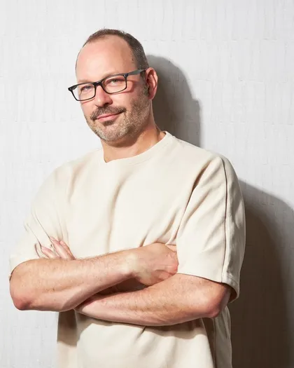 Chris, arms crossed and smiling, in a cream sweatshirt against a white wall.